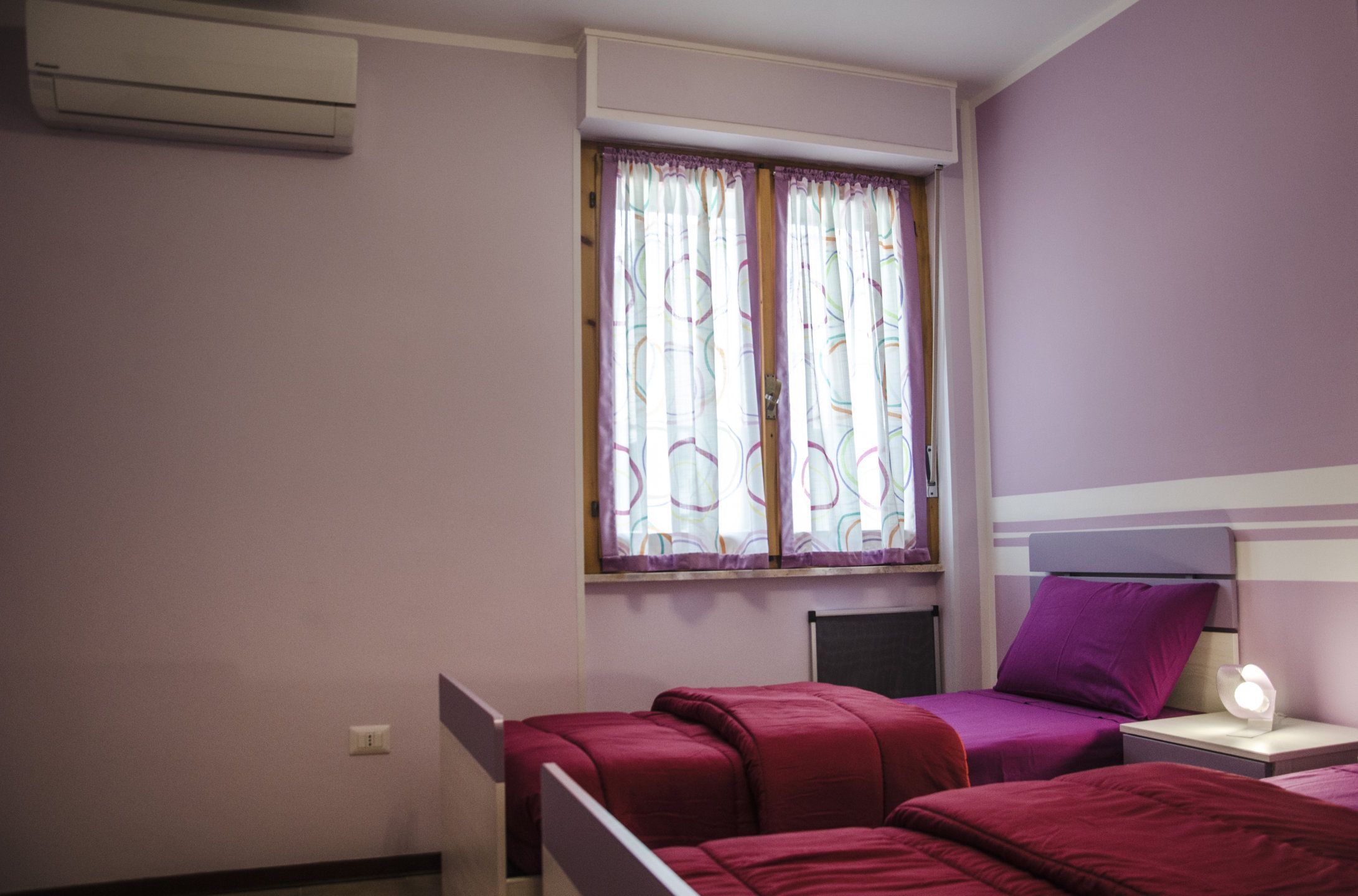 Un dormitorio con dos camas, una ventana con cortinas, una mesita de noche con lámpara y un equipo de aire acondicionado en la pared.