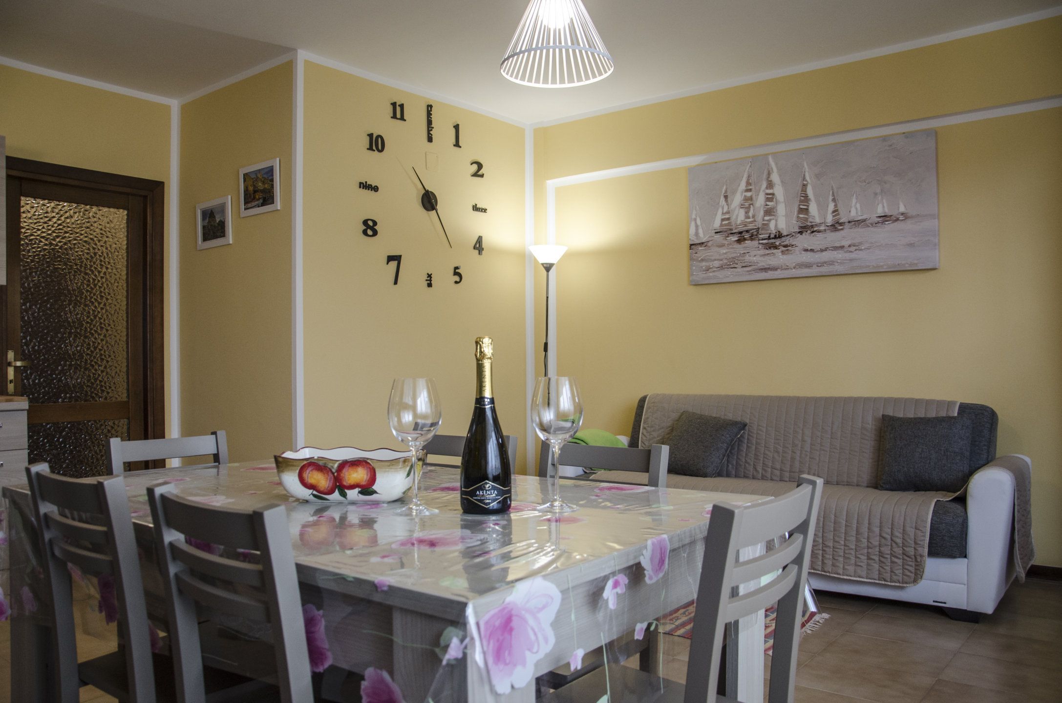 Un comedor con una mesa preparada para dos personas, un reloj en la pared y un cuadro sobre el sofá.