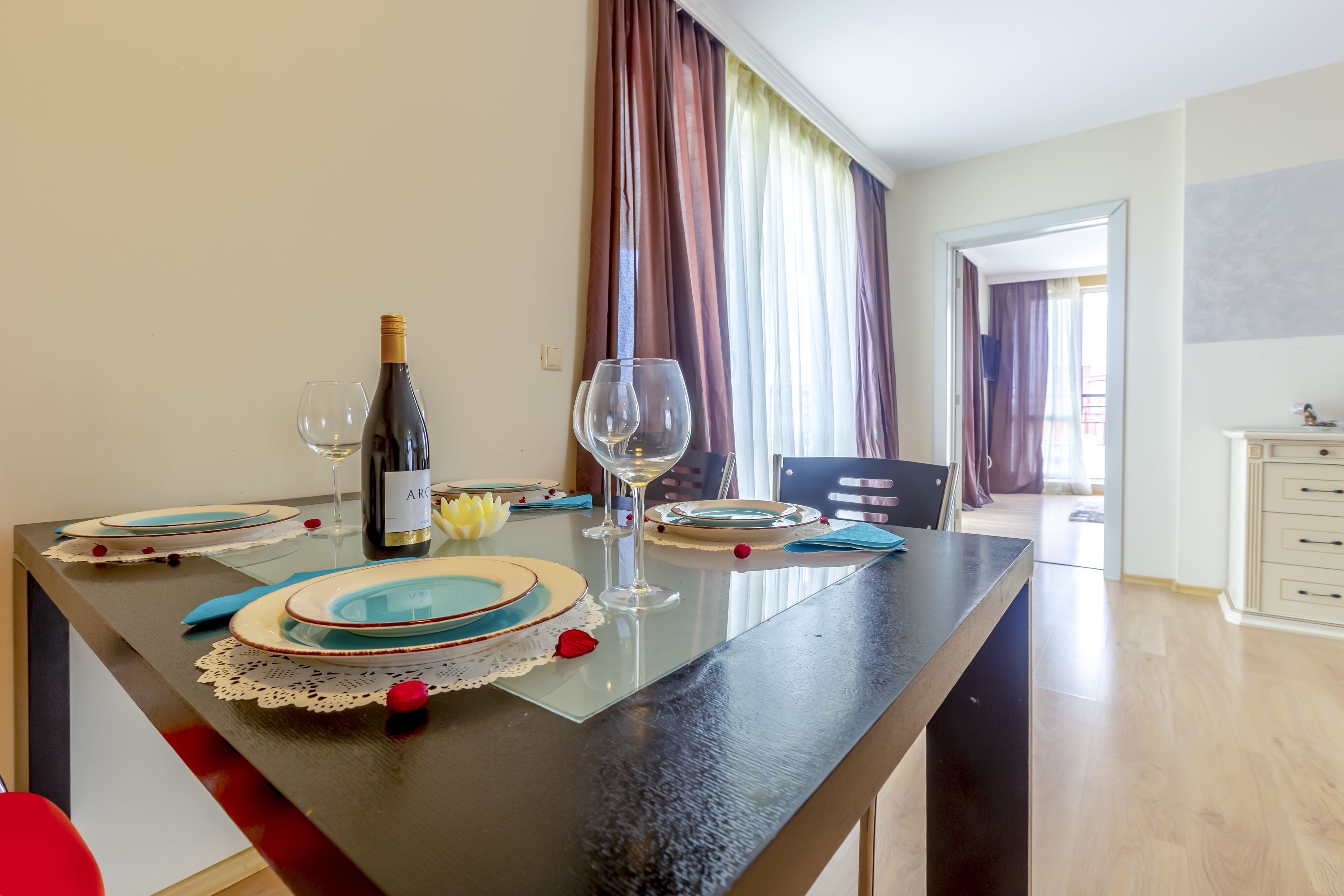 A dining table set with plates, glasses, and a bottle of wine, with a view of a balcony and a door leading to another room.