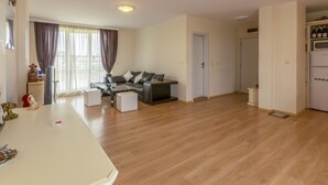 A spacious living area with a sofa, a coffee table, and a dining table. There is a refrigerator and a window with curtains. - Two Bedroom Apartment, Terrace in Aphrodite Palace (Sunny Beach)