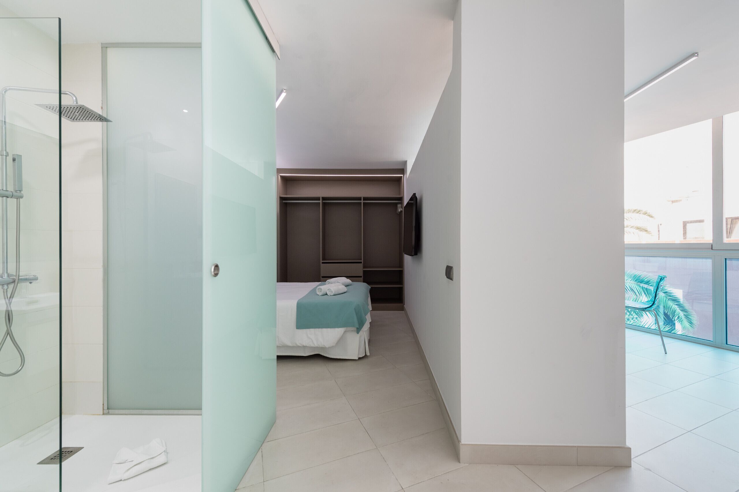 A modern hotel room with a glass shower enclosure, a bed with a teal bedspread, a wardrobe, and a large window offering a view of palm trees.