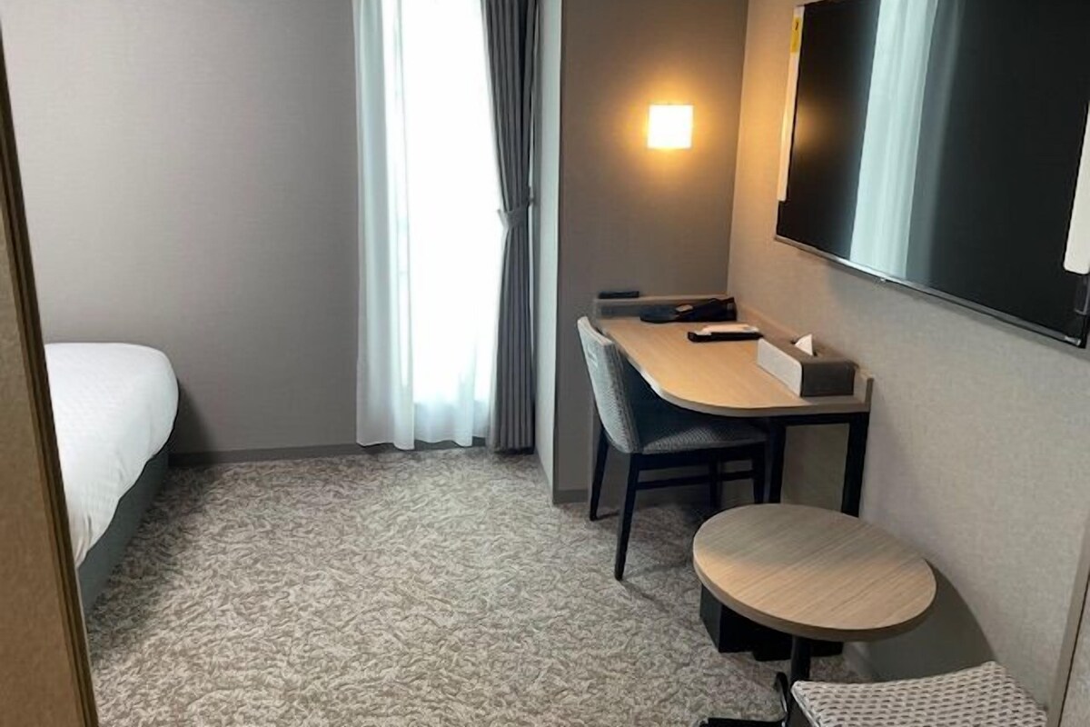 Et hotelværelse med seng, skrivebord, stol, lille bord, fjernsyn og et vindue med gardiner.
