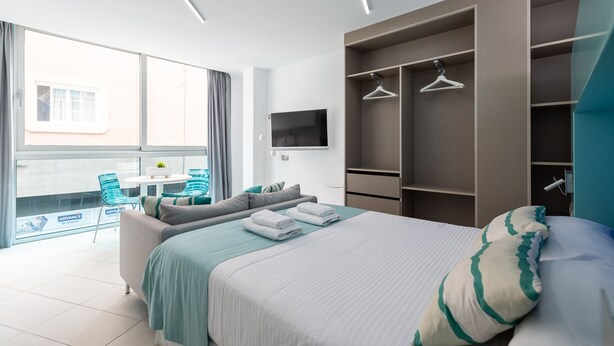 A modern hotel room with a large bed, a TV, a balcony with chairs, and a wardrobe with hangers.