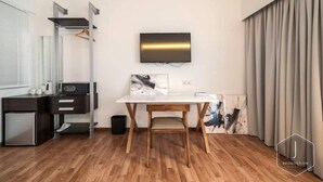 A modern hotel room with a wooden floor, a white desk with a chair, a flat-screen TV mounted on the wall, a small kitchenette with a fridge, and a large window with curtains. - The Boutique Residence Hotel Penang (George Town)