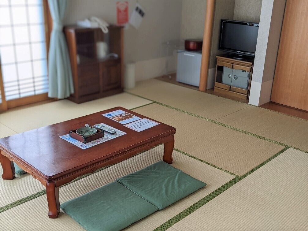 Een traditionele Japanse kamer met tatami vloerbedekking, een lage houten tafel met kussen, een televisie en een kast.