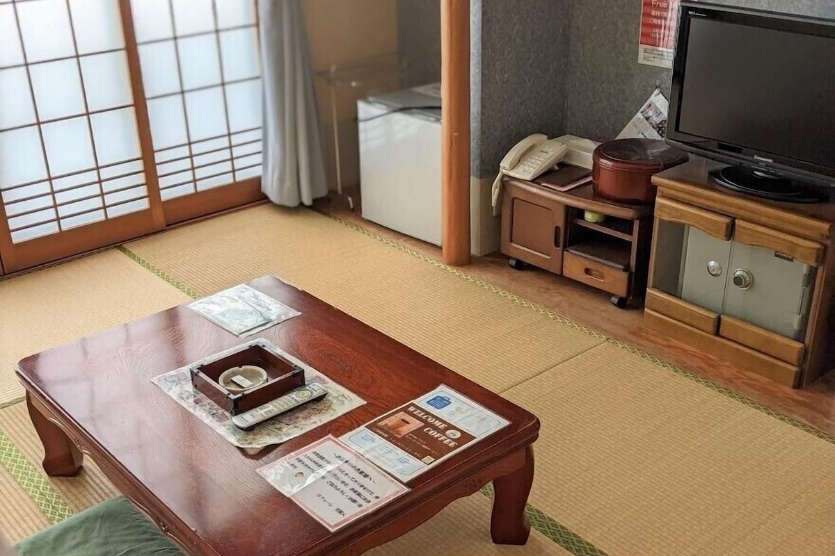 Une pièce de style japonais traditionnel, avec un sol recouvert de tatamis, une table en bois sur laquelle est posé un plateau, un téléviseur et un meuble contenant un coffre-fort.