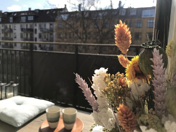 Un balcon donnant sur des immeubles, un coussin, deux tasses et un vase rempli de fleurs.