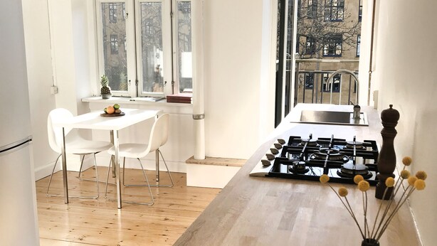 A modern kitchen with a wooden countertop, white chairs, and a view of the city through large windows. - 2 bedroom apartment Vesterbro 1372-1 (Copenhagen)