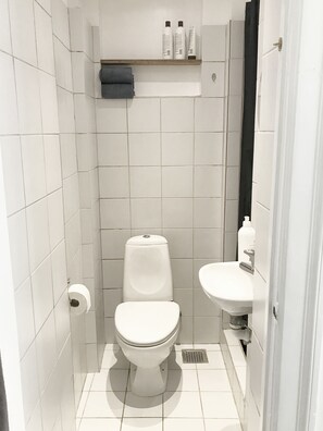 Une petite salle de bain aux murs recouverts de carreaux blancs, équipée de toilettes, d’un lavabo et d’une étagère au-dessus des toilettes.