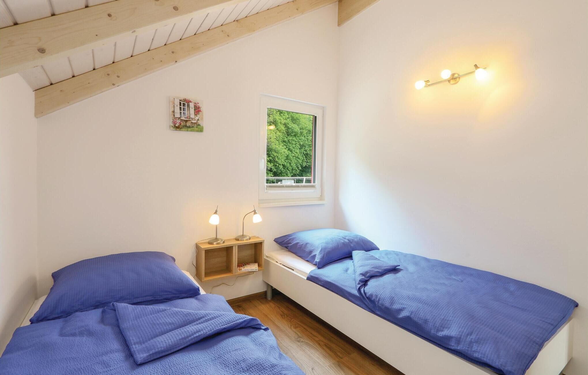 A bedroom with two single beds, a window with a view of greenery, a small bedside table with lamps, and a wall-mounted light fixture.