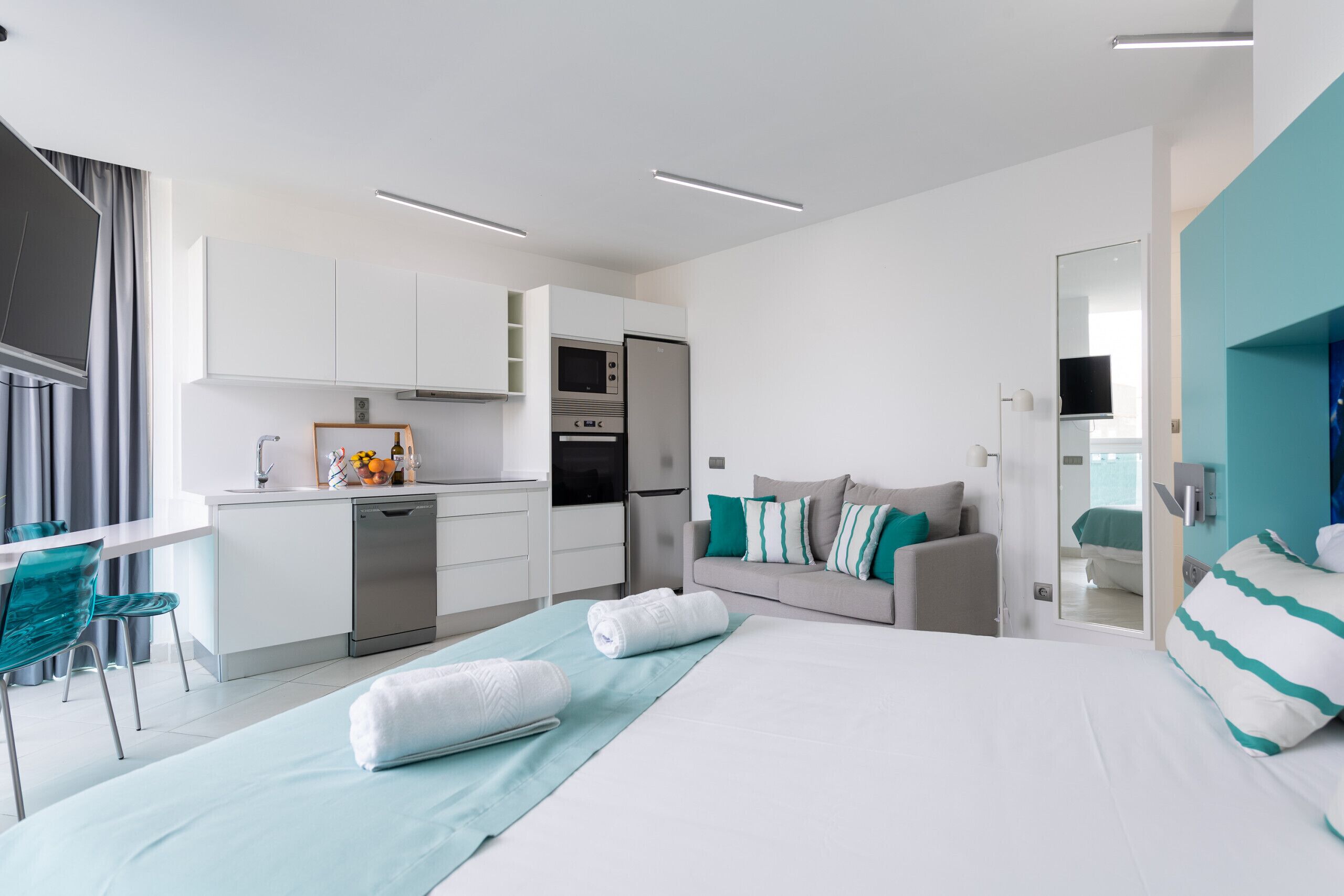 Una habitación de hotel moderna con kitchenette, una cama con ropa de cama blanca y aguamarina, un sofá gris y una silla aguamarina.
