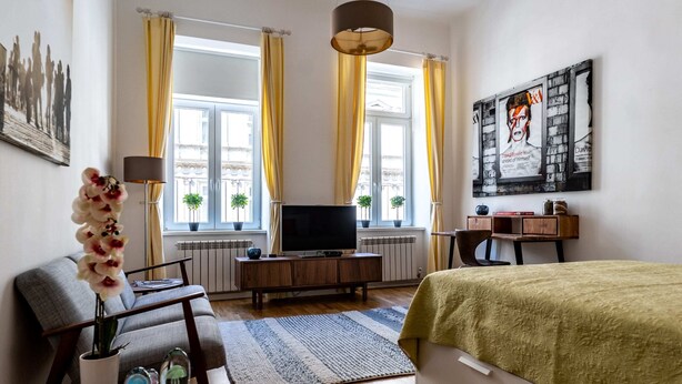 A modern hotel room with a bed, a TV, a sofa, a small table with a vase of flowers, and a large window with curtains. - Boutique Prater Apartment Vienna (Vienna)