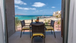 A balcony with wicker chairs and a table, overlooking a beach with a clear view of the ocean and a cityscape in the distance.