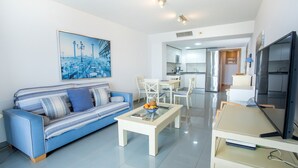 A modern living room with a sofa, coffee table, and a TV. There is a kitchen area with a fridge and a dining table.