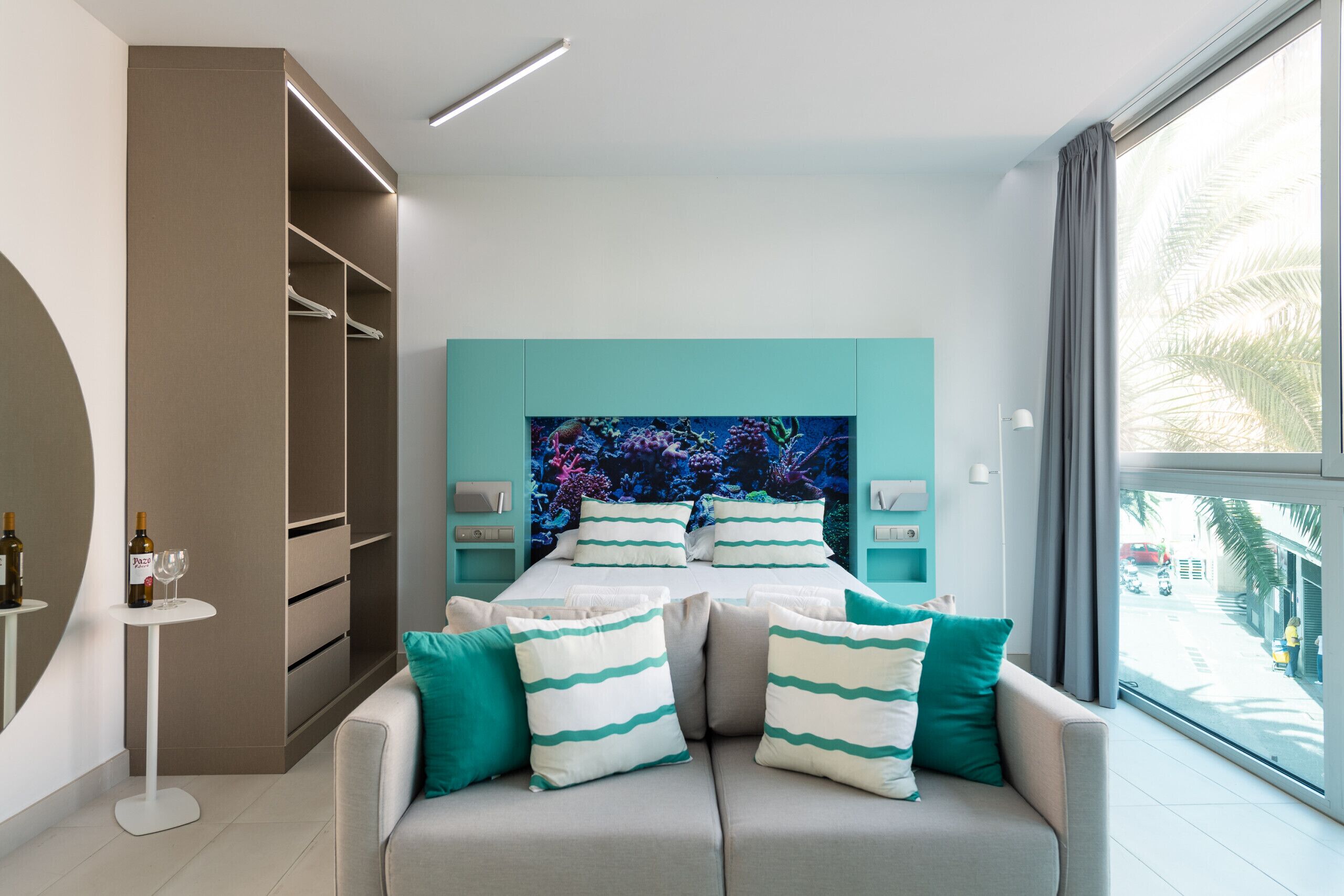 Un dormitorio moderno con cama, paredes de color verde azulado, un ventanal grande y un armario.