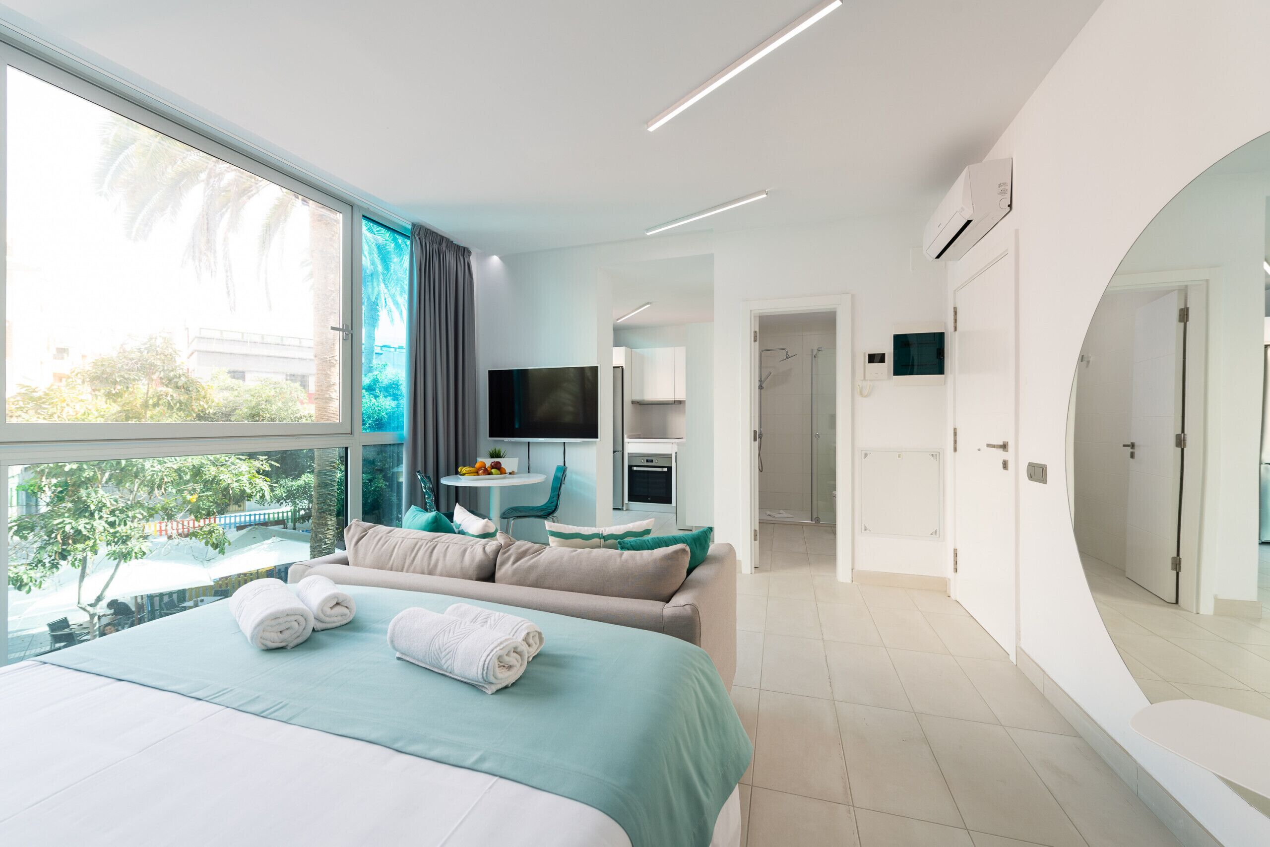 A modern hotel room with a large bed, a TV, a small table with fruit, and a view of palm trees.