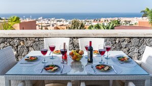 A table set for two with wine glasses, plates of food, and a bottle of wine on a balcony overlooking a coastal city. - Luxury White Villa Sea View, Pool (Adeje)