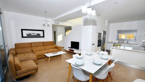 Una sala moderna con un sofá marrón, una mesa de comedor blanca para cuatro personas, y una zona de cocina con refrigerador y microondas.