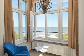 A room with a large window overlooking the sea, a blue chair, a small white table, and a hanging light fixture. -  Strand- und Wellnesshotel Preussenhof (Zinnowitz)