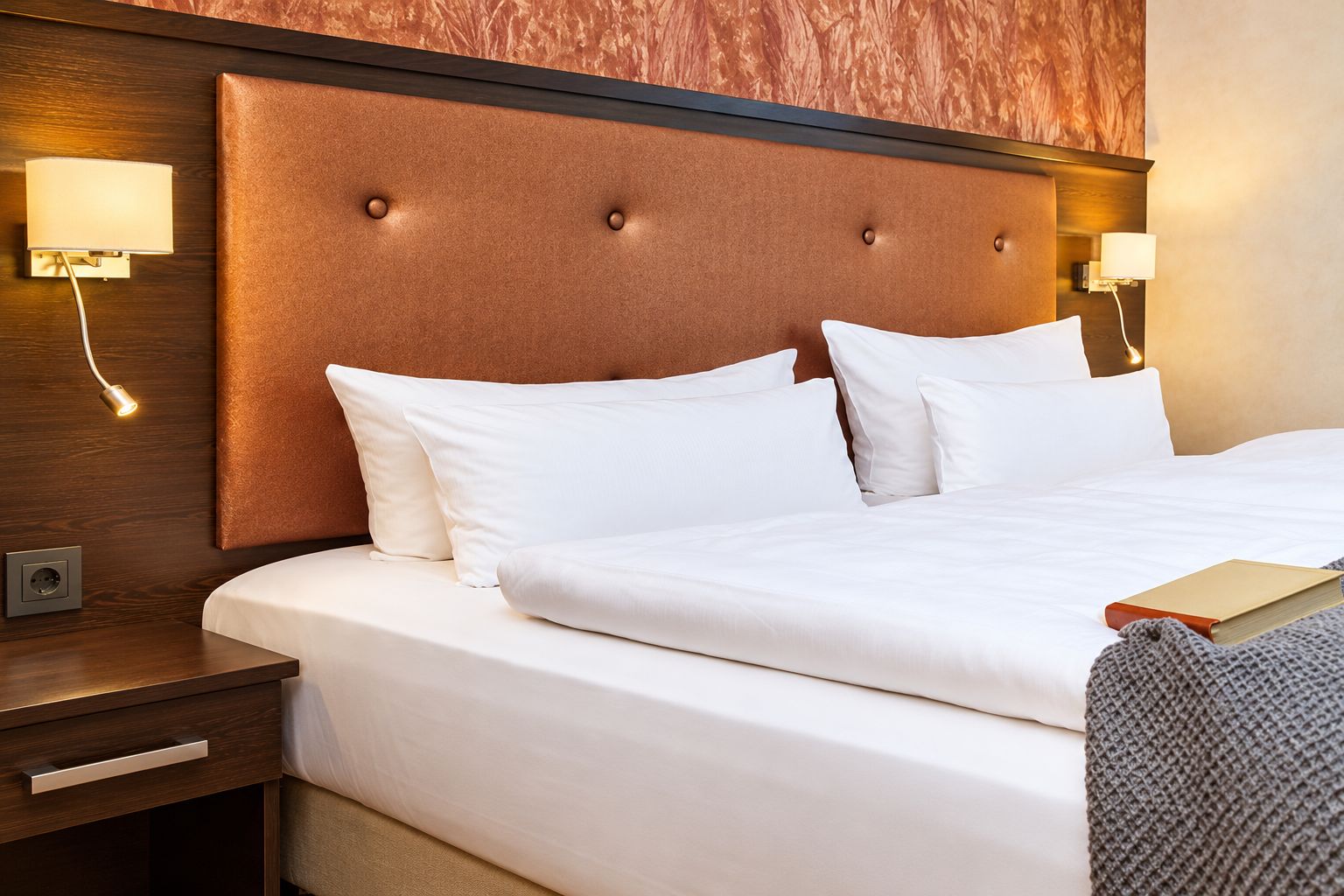 Habitación de hotel con una cama grande, dos lámparas de noche, una mesita de noche y un libro sobre la cama.