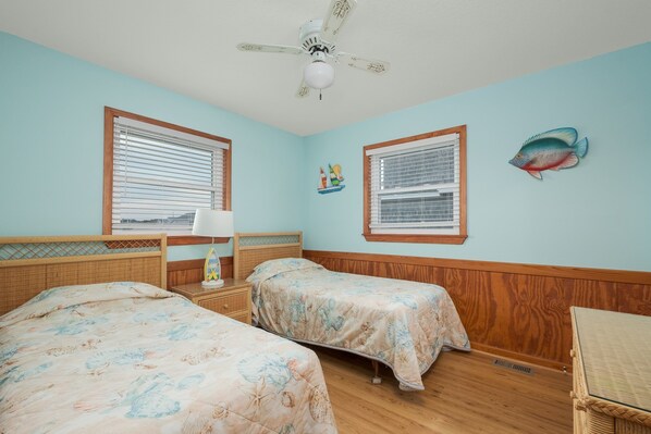 A room with two beds, a ceiling fan, and a window with a sailboat decoration.