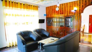 Una habitación con panel de madera en la pared, ventilador de techo, sofá, dos sillones y una mesita con mantel floral.