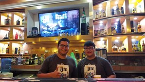 Two individuals standing in front of a bar with shelves stocked with various liquor bottles. A television is mounted above the bar, displaying a scene with a person. - Hotel Club Quinta Pat Resort & Restaurant (San Antonio de Oriente)