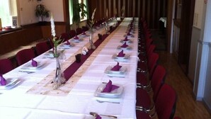 Una larga mesa de banquete dispuesta con manteles blancos, servilletas moradas y cubiertos, flanqueada por sillas rojas en una habitación con paneles de madera y un ventanal grande.