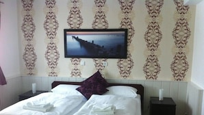 Una cama bien hecha con sábanas blancas y una almohada morada, una mesita de noche pequeña con una lámpara, y un televisor de pared mostrando un paisaje urbano.
