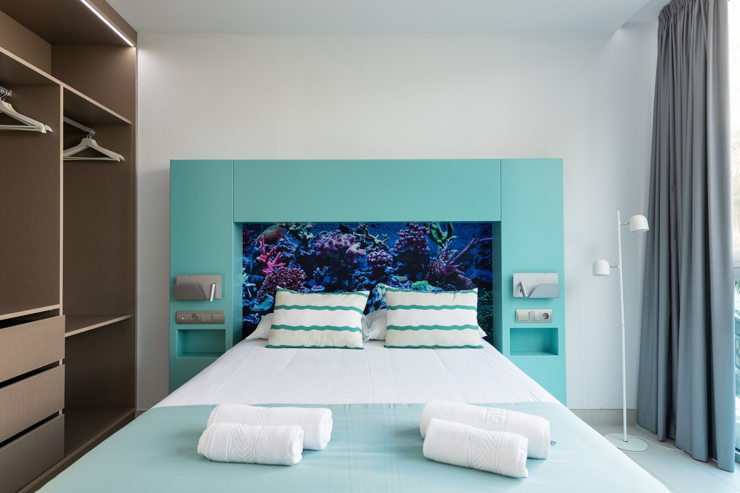 A modern hotel room with a teal-colored bed, white bedding, and a coral reef mural on the headboard. There are two bedside tables with lamps, a wardrobe, and a large window with curtains.