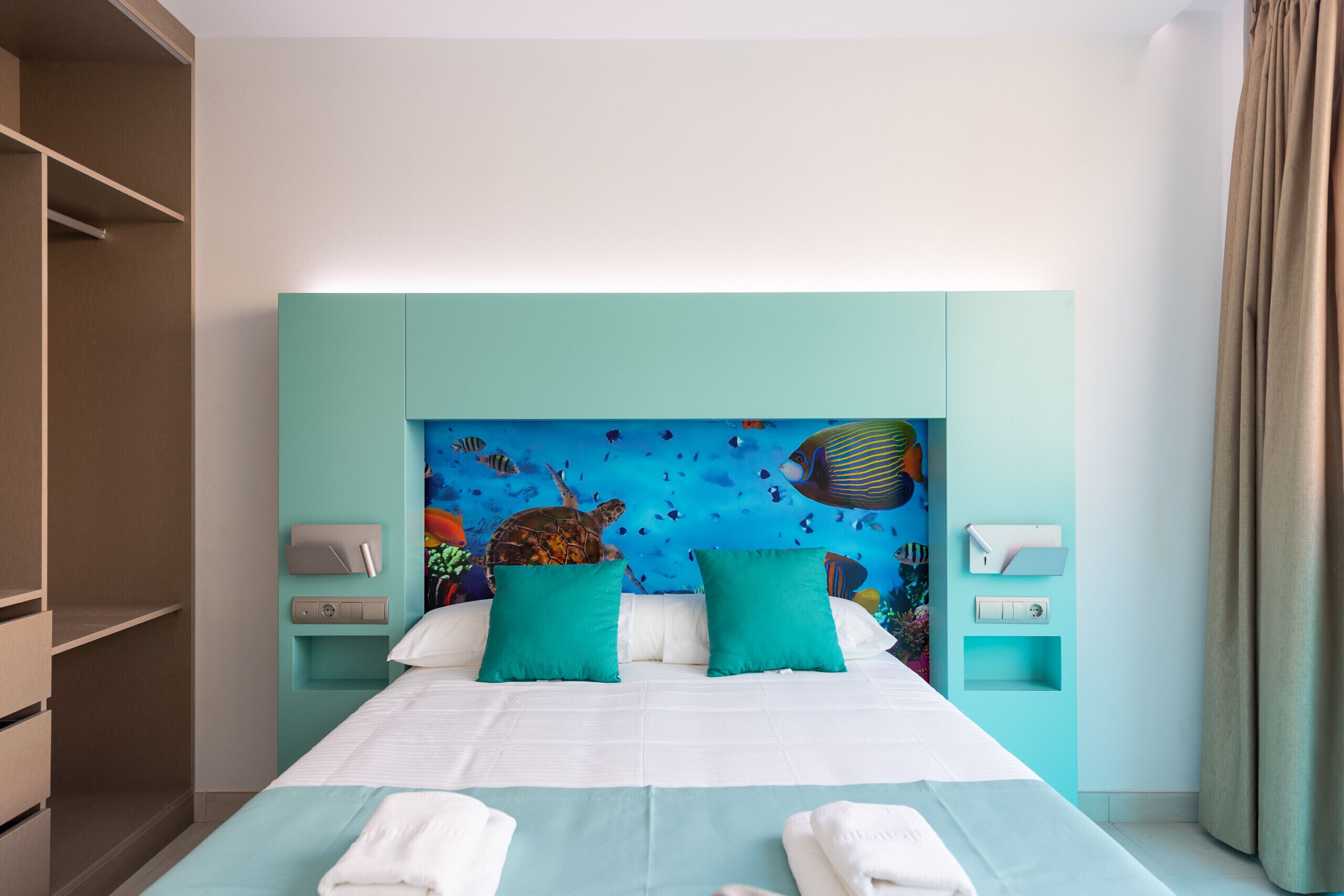 A hotel room with a bed, teal pillows, a mural of underwater scene, and a wooden wardrobe.