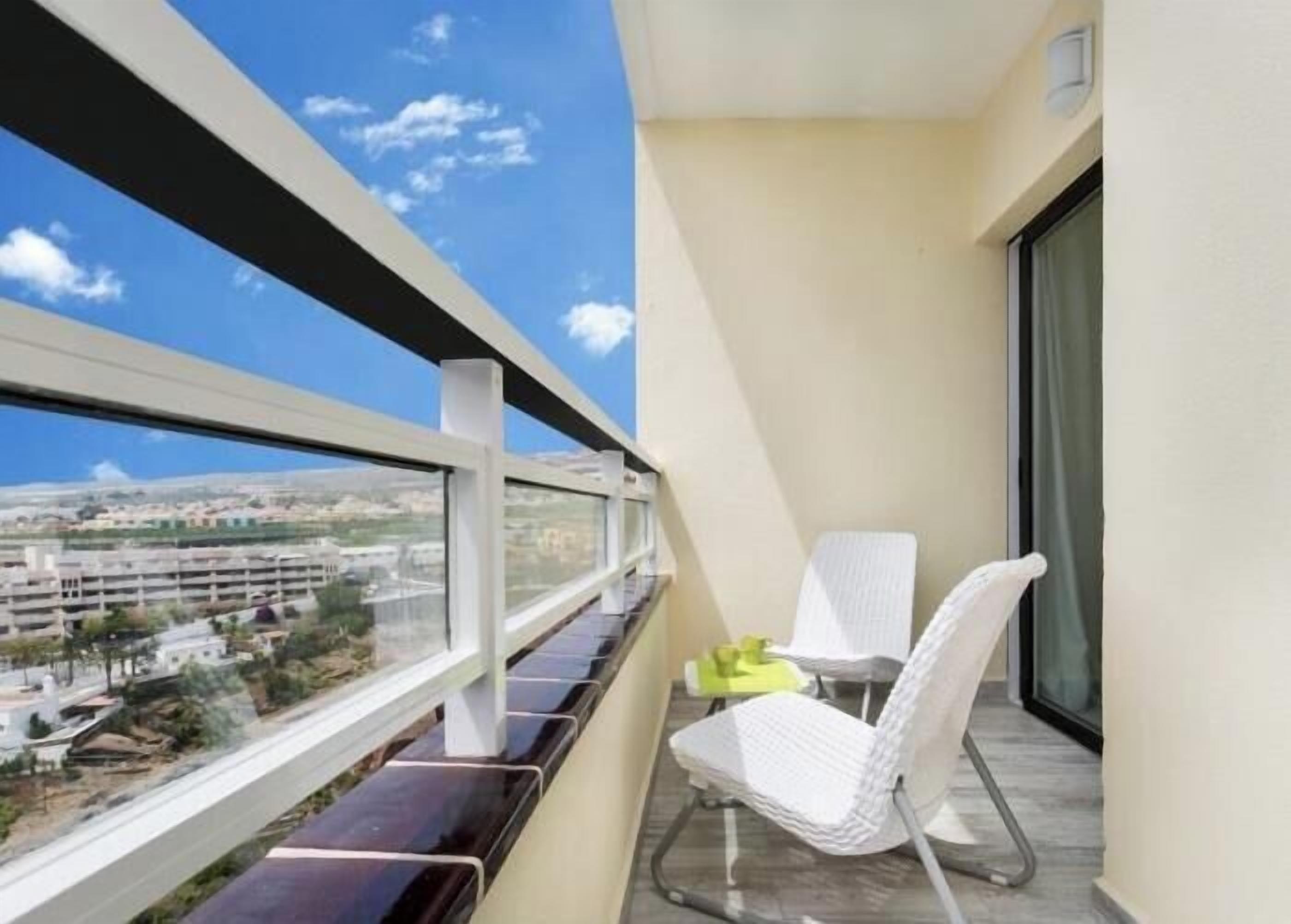 Un balcon donnant sur une vue de la ville, une chaise blanche et une petite table sur laquelle se trouve un objet jaune.