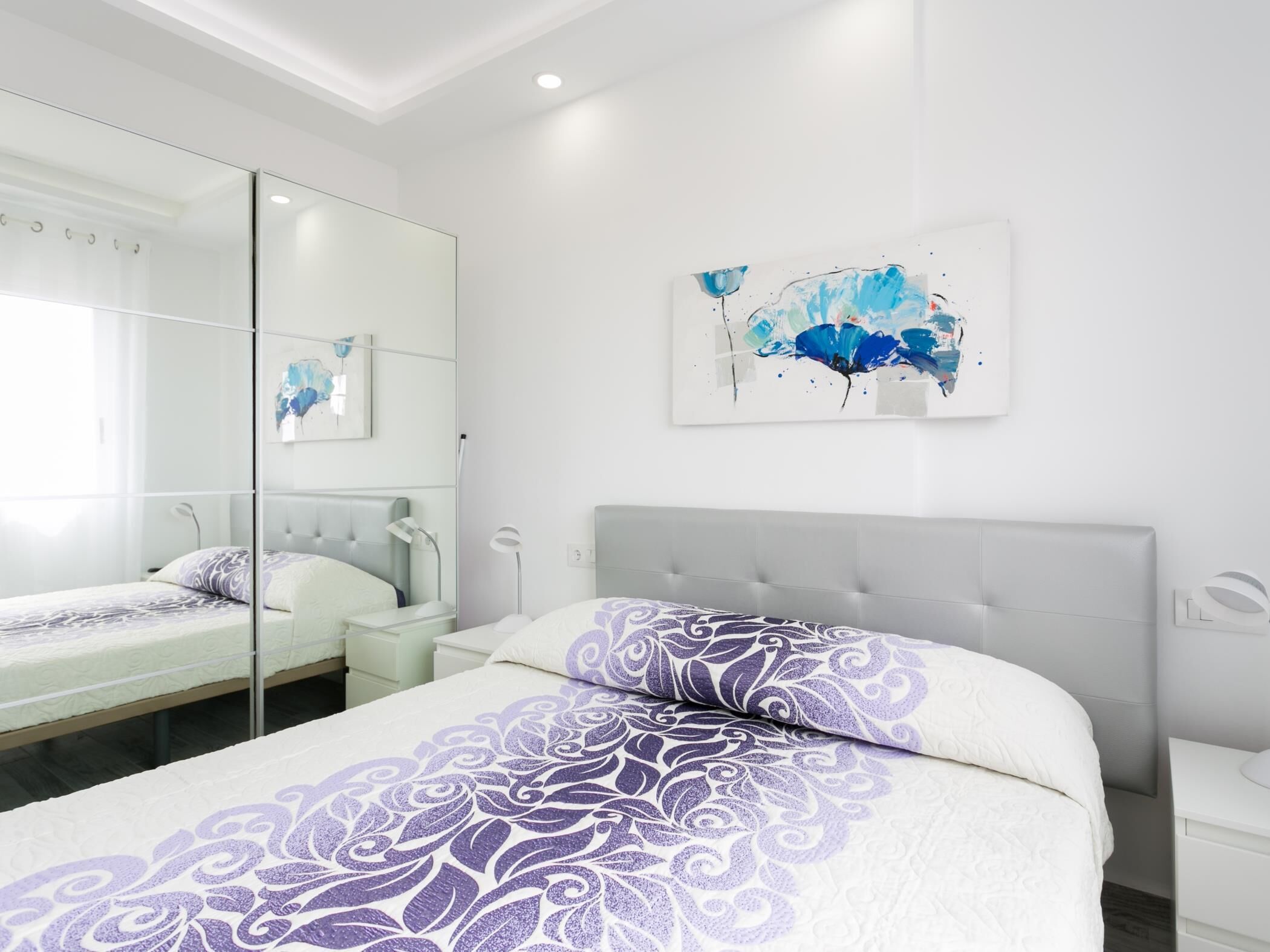 A modern bedroom with a large bed, a bedside table, a nightstand, a mirror, and a wall-mounted abstract painting.