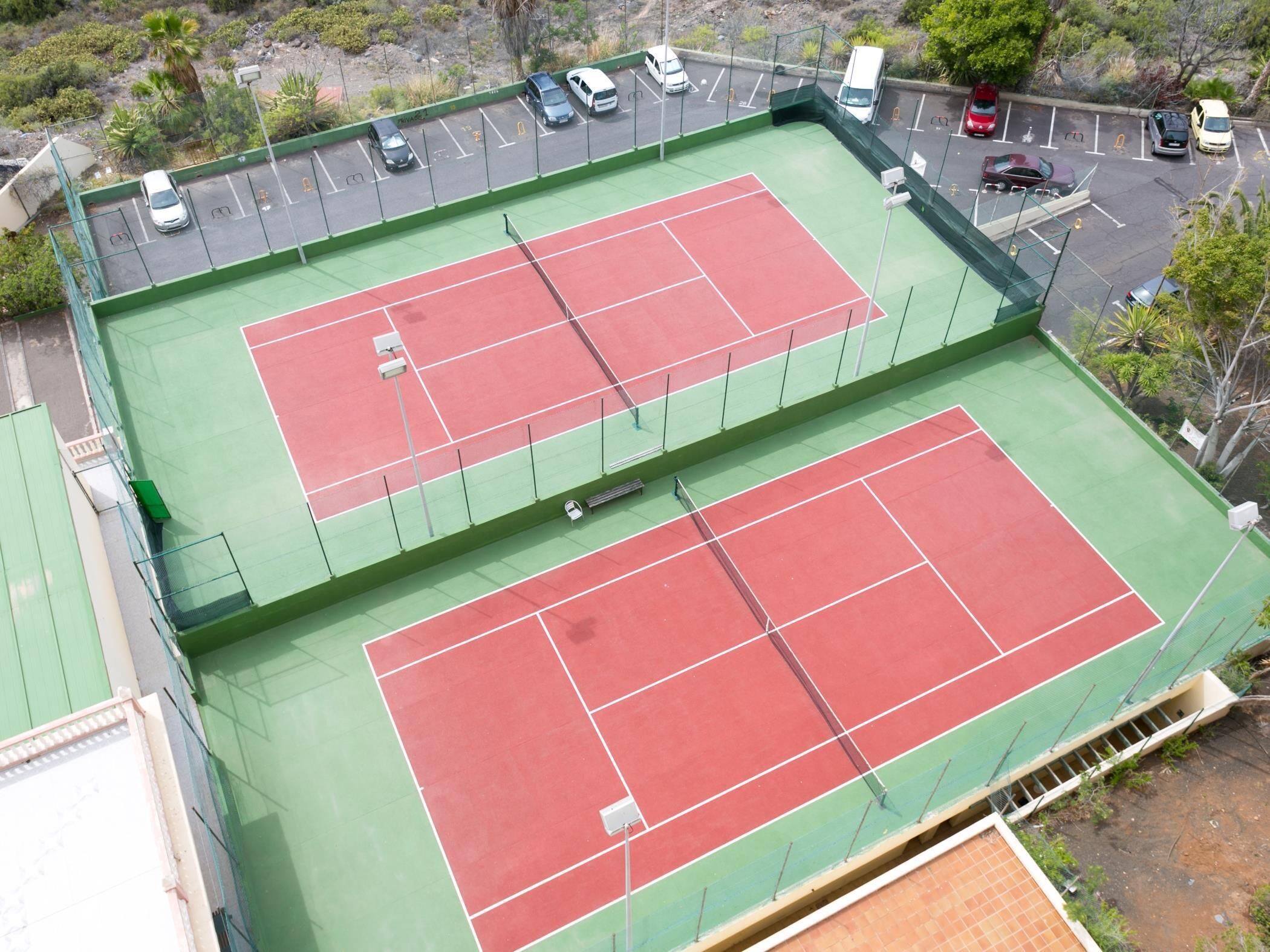 An outdoor tennis court with a green and red surface, surrounded by a parking area with several cars.