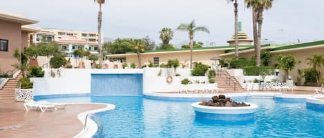 A swimming pool with a waterfall feature, surrounded by palm trees and lounge chairs.