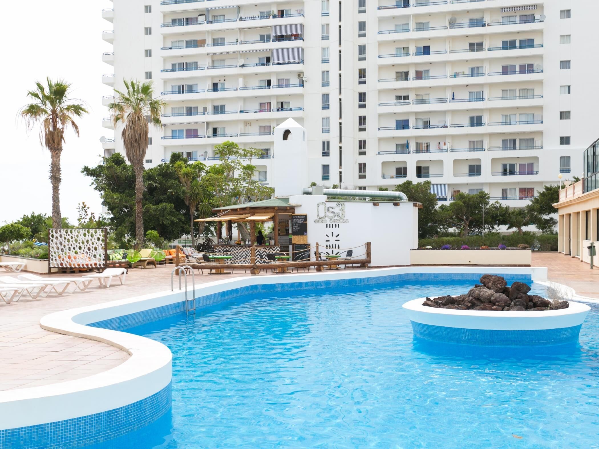 A swimming pool with a lounge area, palm trees, and a high-rise hotel in the background.
