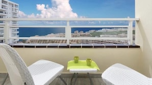 A balcony with white wicker furniture, a small table with two green cups, and a view of the sea and buildings.