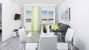 A modern living room with a glass dining table, white chairs, a grey sofa with green cushions, a flat-screen TV mounted on the wall, and a large window with a view of the sea.