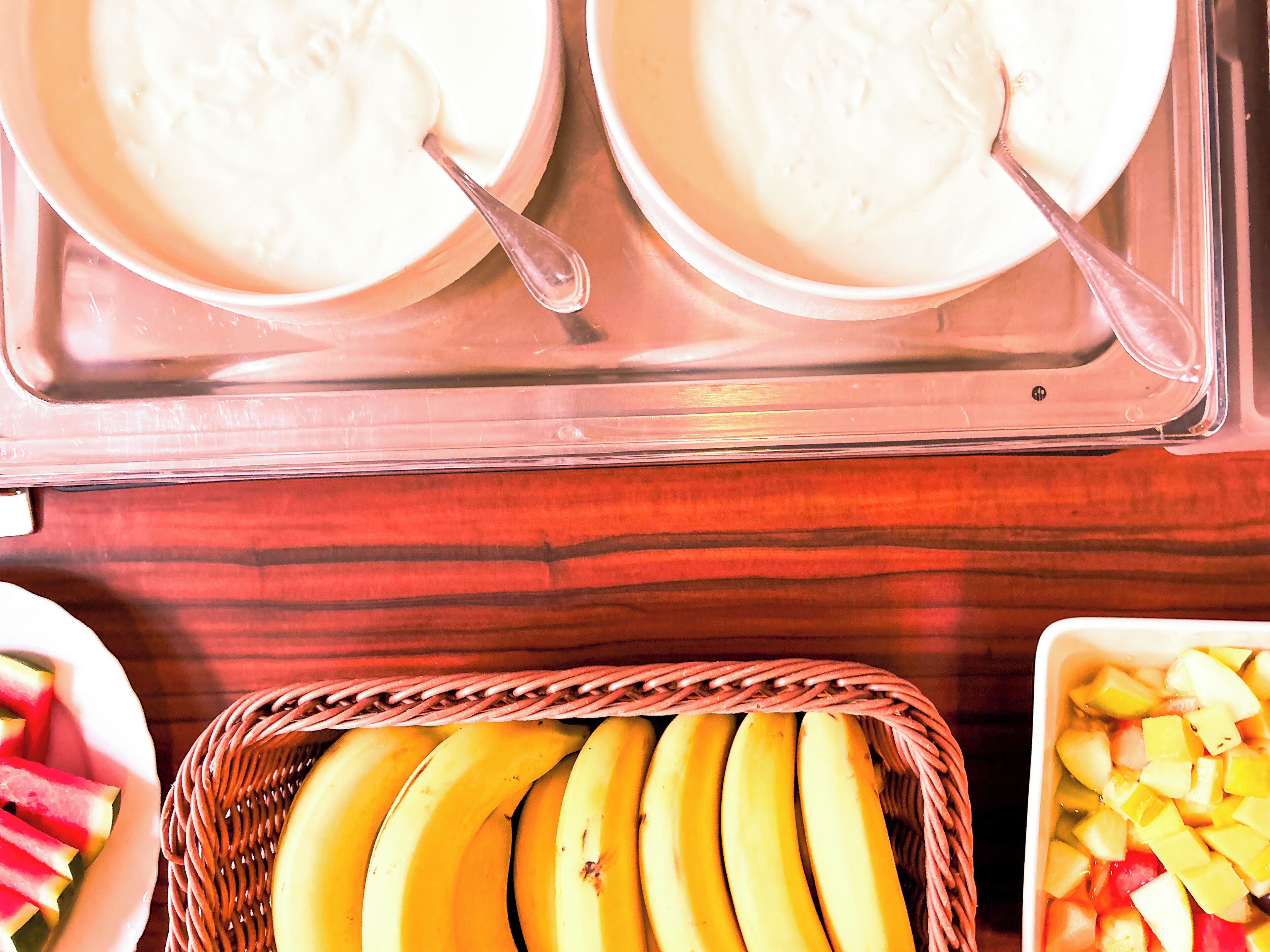 Une table de buffet avec des plats contenant des aliments blancs, un panier de bananes et un saladier de fruits coupés en dés.