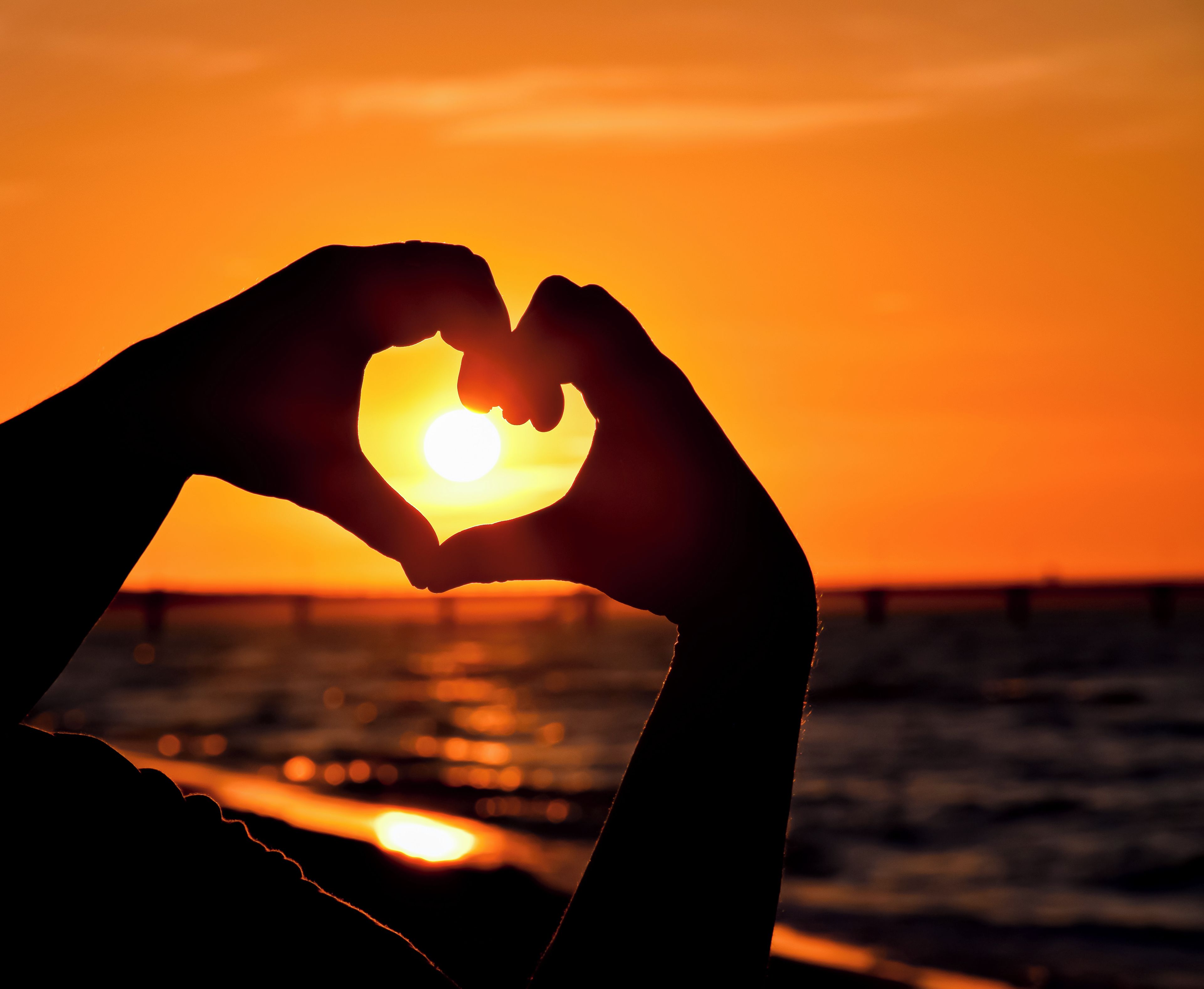 Manos formando un corazón, con el sol en el centro, siluetas contra un atardecer.