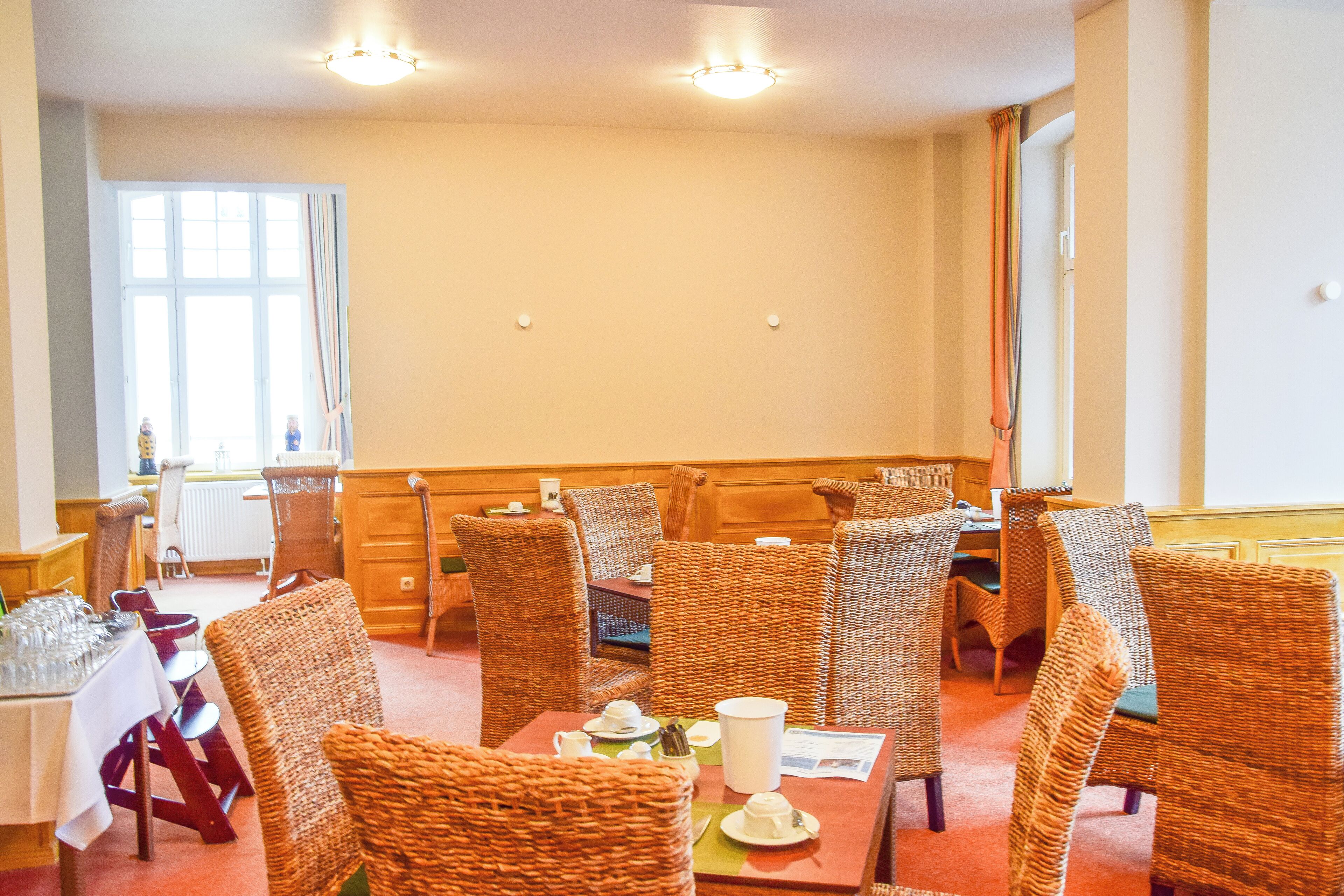 A dining area with wicker chairs and tables, a window with curtains, and a radiator.