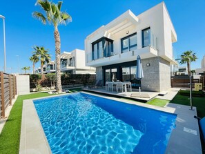 Una casa moderna de dos pisos con piscina, mobiliario exterior y palmeras.