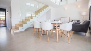 Ein modernes Wohnzimmer mit einer Glaskonstruktion als Treppe, einem Essbereich mit weißen Stühlen und einem grauen Sofa.