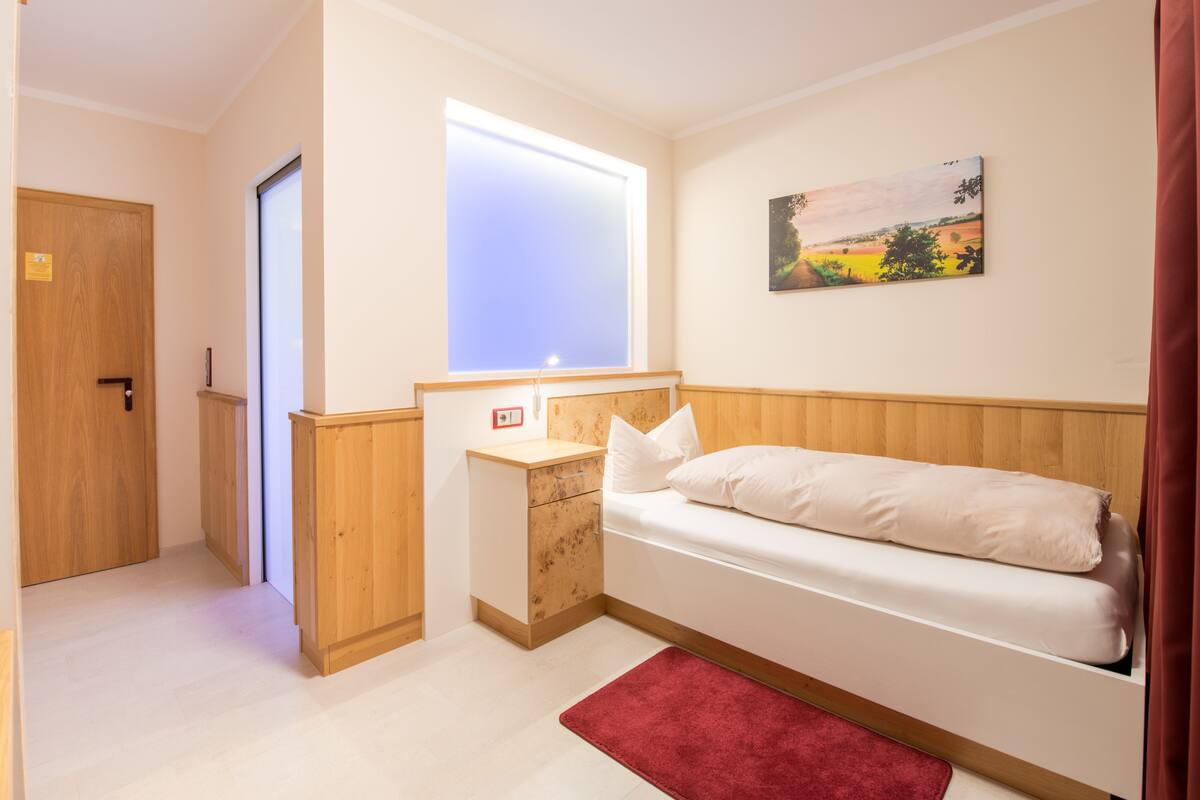 Une petite chambre à coucher, bien rangée, avec un lit simple, une table de chevet, une fenêtre avec des stores, une porte et un tableau au mur.