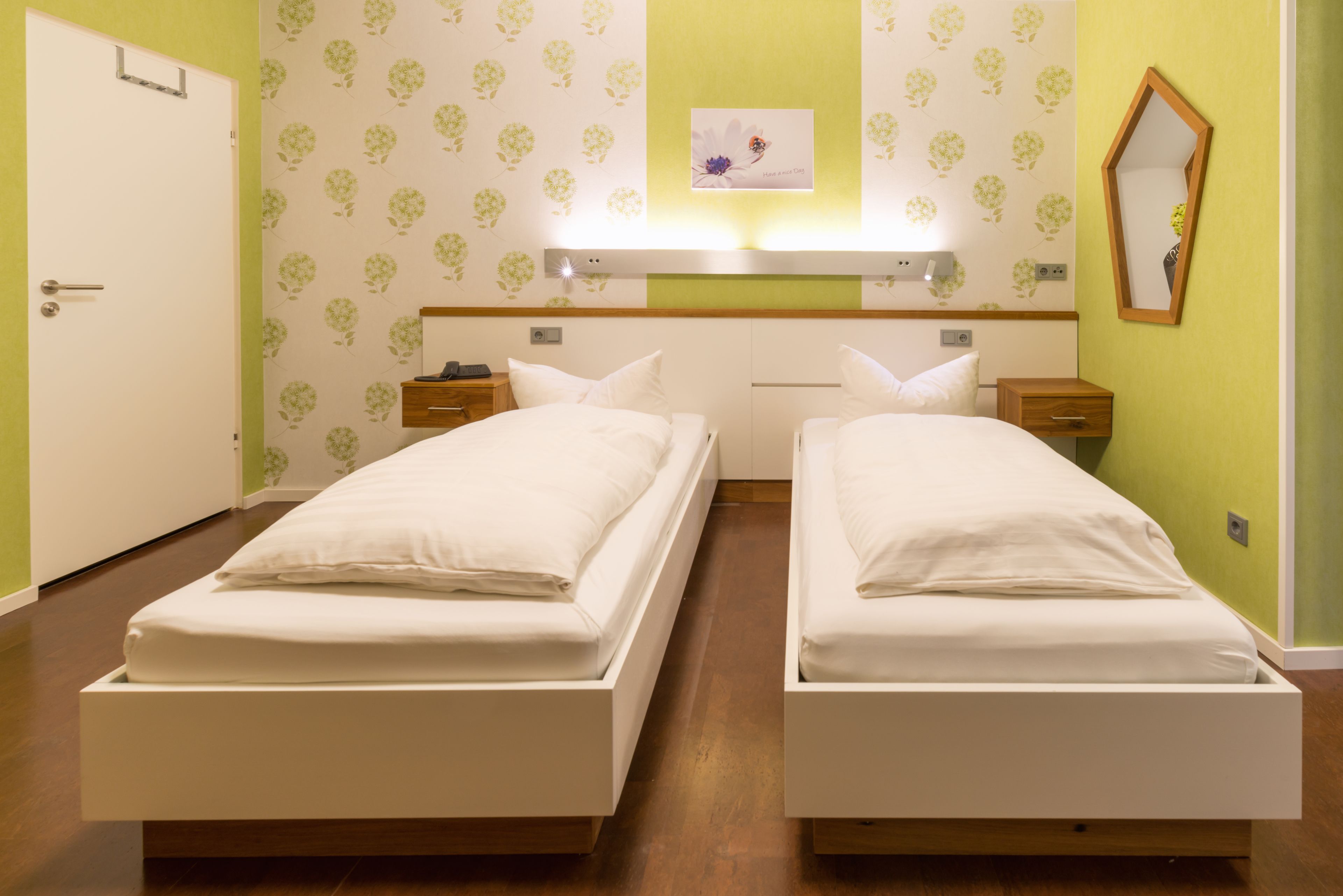 A hotel room with two single beds, a nightstand with a lamp, a mirror, and a wall-mounted light fixture.
