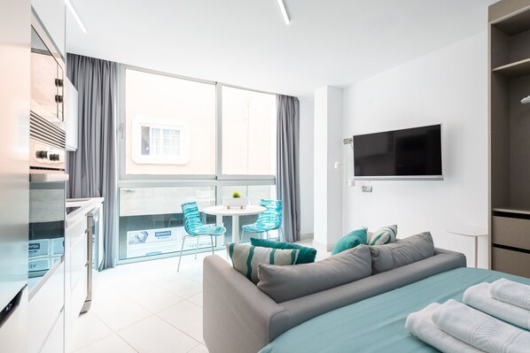 Habitación de hotel moderna con una cama grande, televisión montada en la pared, balcón con mesa y sillas, y vistas a la ciudad.