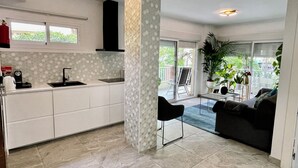 A modern kitchen with white cabinets, a black sink, and a tiled floor. There is a black chair and a gray sofa in the living area.