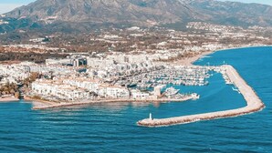 An aerial view of a coastal town with a marina, buildings, and a mountain in the background.