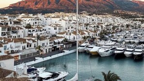 A coastal town with white buildings, a marina with numerous boats, and a mountain in the background.