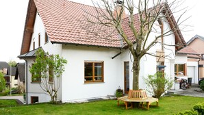 Ett vitt hus med ett rött tegeltak, en liten trädgård med en bänk och ett träd framför.